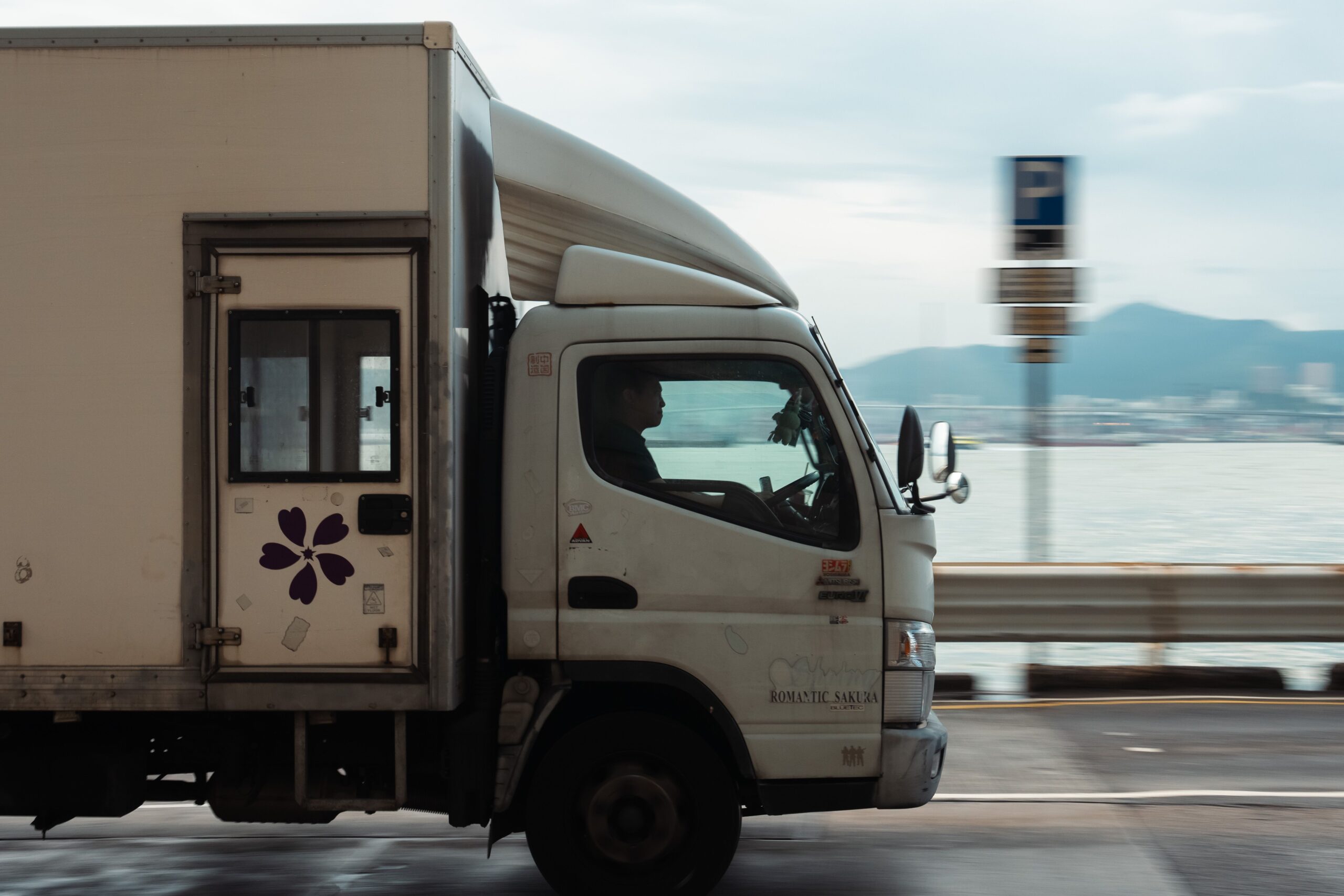 A delivery truck driving along a coastal road with a mountain backdrop on a clear day.